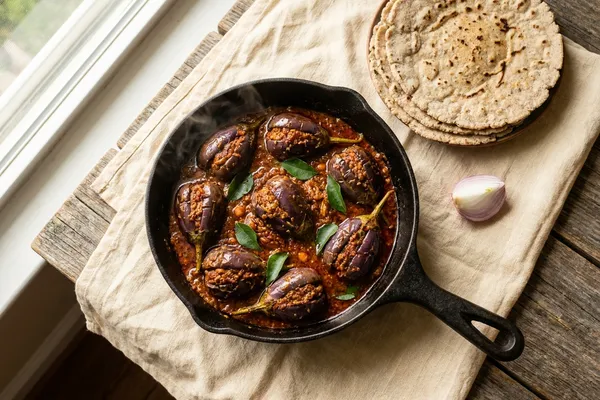 Four small purple brinjals split into quarters and packed with a deep red-brown peanut-and-sesame masala, simmering in a glossy gravy in a black cast-iron pan, garnished with curry leaves.