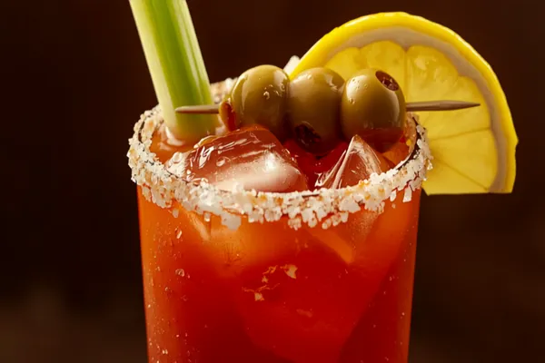 A tall Bloody Mary over ice with a celery-salt rim, garnished with a celery stick, olives and a lemon wedge.