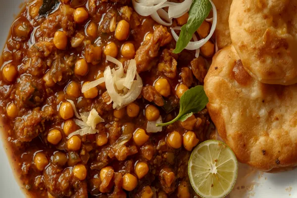 A plate of dark, spiced chickpea chole topped with ginger and onion beside two large puffed golden bhature, with sliced onion, green chili, and lime.
