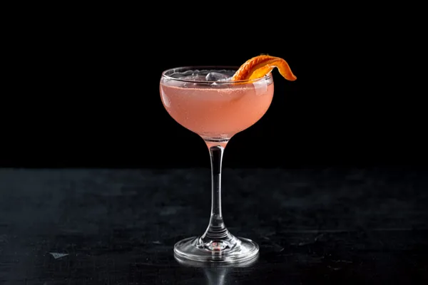 A pale pink Cosmopolitan served up in a chilled coupe with a flamed orange twist, sleek dark bar and soft highlights.