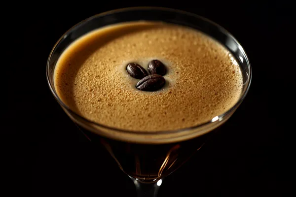 An espresso martini in a chilled coupe with a thick glossy crema foam on top and three coffee beans floating, dark and elegant on a bar.