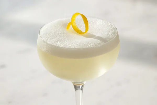 A gin sour in a coupe with a silky foam cap and a lemon peel, crisp bright light.