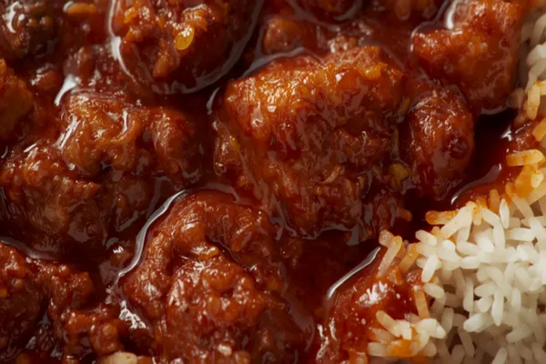 A deep red Goan pork vindaloo in a bowl, tender chunks of pork in a thick glossy chili-vinegar gravy with oil glistening on top, beside white rice.