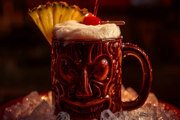 A Jungle Bird cocktail in a tiki mug over crushed ice with a frothy top, pineapple wedge and cherry, moody tropical light.