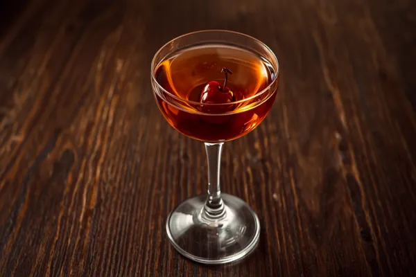 A deep amber Manhattan served up in a coupe glass with a brandied cherry, on a dark wood bar with warm low light.