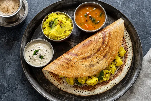 A long, golden, crisp masala dosa folded over spiced yellow potato on a steel plate, with small bowls of white coconut chutney and orange sambar alongside.