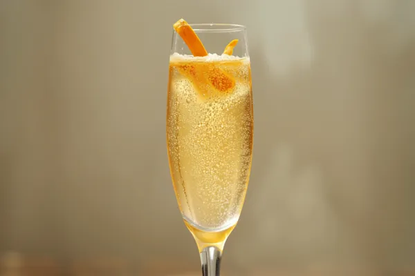 A Mimosa in a champagne flute, pale golden-orange and gently fizzing, with a thin orange twist.