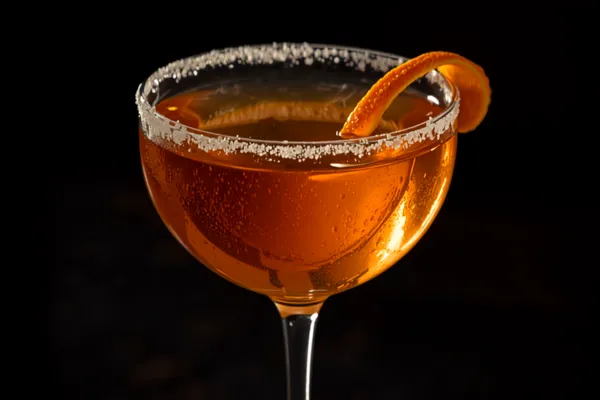 A Sidecar straight up in a coupe with a half sugar rim and an orange twist, amber and bright on a dark bar.