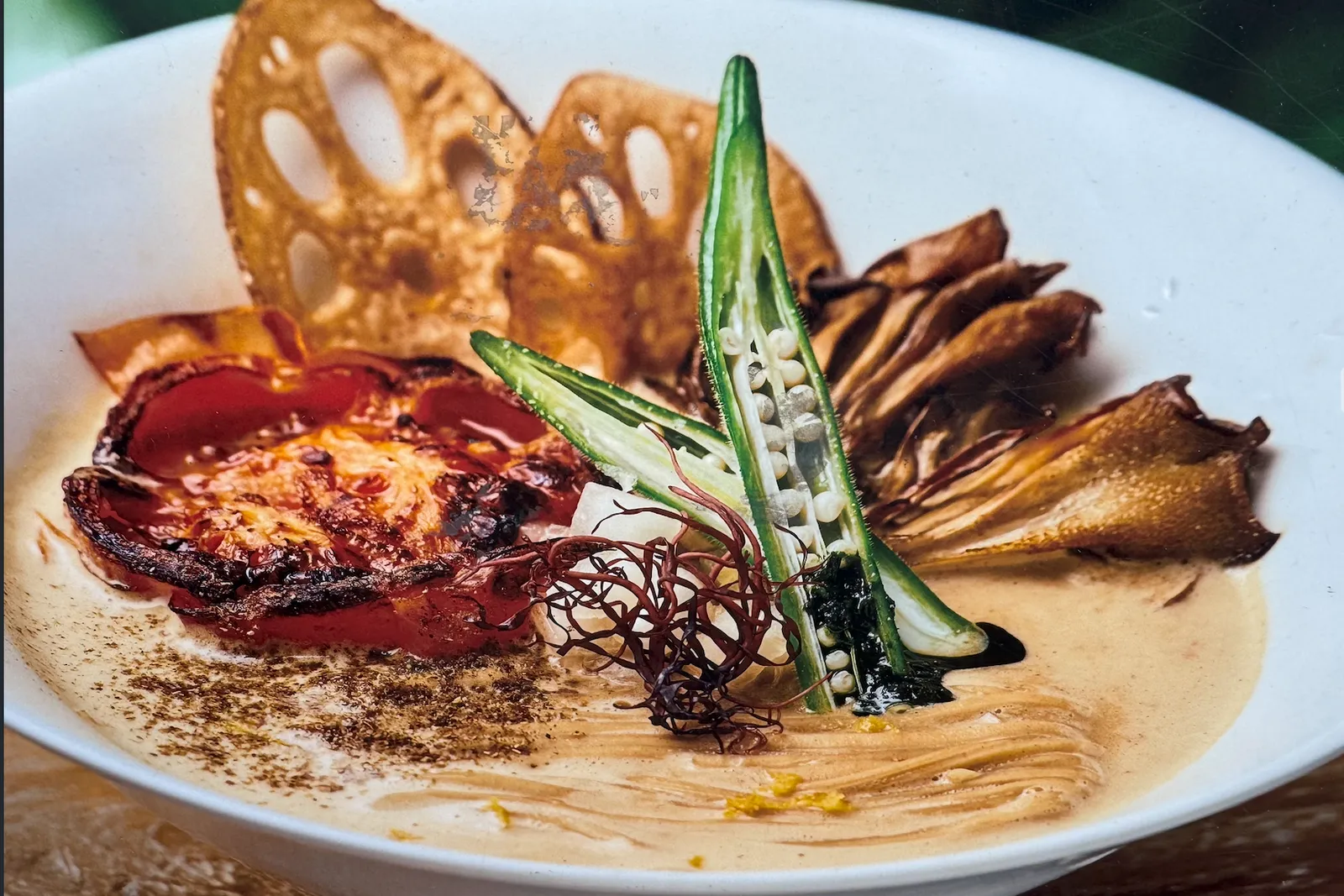 A wide white bowl of spicy vegan miso ramen with a pale, creamy, frothed sesame-oat broth. Toppings are composed in distinct zones: a blistered charred tomato half at the left, two crisp lacy lotus-root chips standing behind, a large fanned cluster of roasted maitake mushroom at the right, a bright-green okra pod split lengthwise to show its seeds standing upright in the centre, a small nest of fine red-brown thread chili, dark greens, a dusting of smoky spice fanned across the near edge of the broth, flecks of yellow yuzu zest, and a ribbon of dark chili oil — on a wooden table.