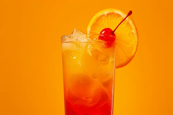 A tall Tequila Sunrise over ice with a red grenadine gradient rising into golden orange juice, orange wheel and cherry.