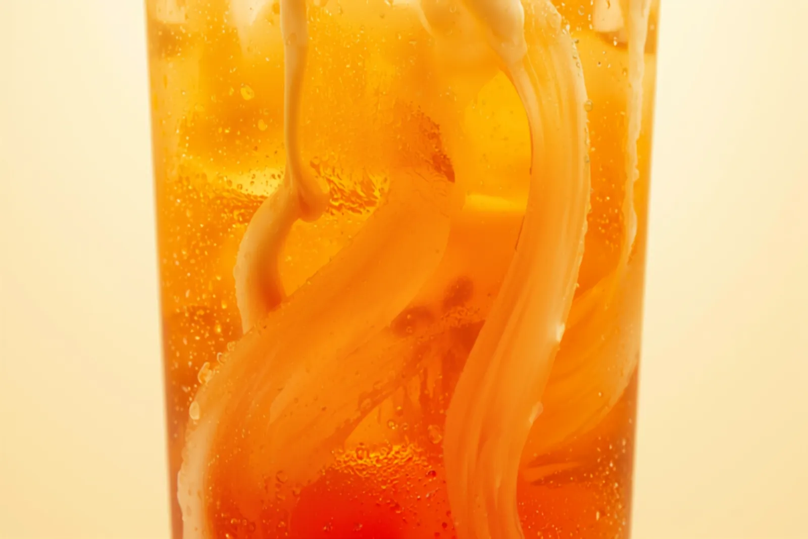 A tall glass of bright orange Thai iced tea over ice with a swirl of cream floating on top and being stirred down, condensation on the glass.