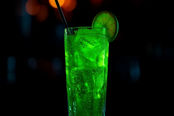 A tall neon-green Tokyo Tea over ice with a lime wheel and a straw, bright and glowing against a dark bar.